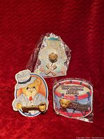 Three vintage Cherished Teddies brooch pins displayed on red fabric background. Two pins have original card backing and one is wrapped in plastic.