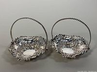 Pair of ornate sterling silver repousse reticulated bowls with twisted handles on a flat surface.
