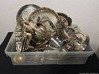 Plastic bin filled with assorted vintage silver plated serving pieces such as trays, teapots, and bowls showing patina and tarnish.