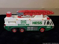 Side profile of 1996 Hess fire truck toy showing six wheels, white and green body, red ladder and siren light, labeled 'Hess Gasoline'.
