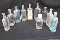 Lot image showing 10 antique and vintage glass bottles of various shapes and sizes, mostly clear glass with some light blue tinting, one bottle labeled, placed on black background.