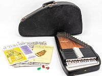 Full shot of the Oscar Schmidt OS-15B autoharp with its black hard carrying case, music books, finger picks, and a green plastic pick placed alongside.