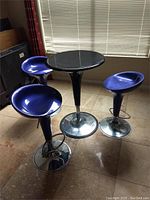 The lot showing one small black round pub table and three blue half moon stools with chrome bases arranged in a room corner.