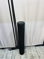 Front-facing photo showing cylindrical black textured foam roller standing upright on carpeted floor with white curtain backdrop.