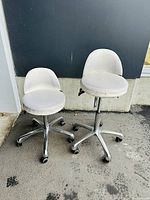Two white cushioned stools with silver metal bases side by side, taller with backrest, shorter without backrest