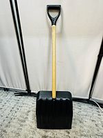Front view of a snow shovel with a black plastic blade and wooden handle with a black plastic D-grip.
