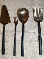 Four large bronze serving utensils with black hammered handles on a tablecloth: a cake/pie server (triangular flat head), small fork with three tines, large spoon, and large fork with four tines.
