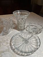 Four clear crystal decorative items on a table with a beige tablecloth: a vase, a boot-shaped figurine, a bowl, and a small platter, showing overall appearance and condition.