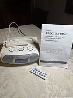 Electrohome dual alarm clock radio with universal dock for iPod 5, remote control and owner's manual displayed on table.