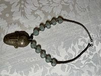 Top view of the bronze tone mask pendant necklace showing the mask face and greenish-white glass beads on bronze tone chain laid on a white patterned fabric.