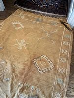 Full image of larger rug laid on floor showing overall condition and border designs with light blue and cream embroidery on tan base.