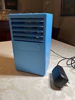 Front view of blue personal air cooler showing air vents and black power adapter with cord placed on table.