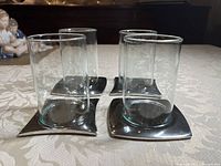 Four clear glass cylindrical votive candle holders each sitting on a square silver tone base with curved edges. Items placed on beige patterned cloth.