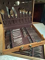 Top view of teak display box open showing handles and arranged bronze cutlery including forks, spoons, and knives in designated slots.