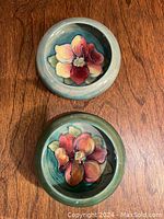 Two round Moorecroft bowls with raised floral designs and green-blue backgrounds on a wooden surface.