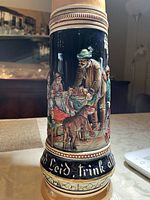 Full view of the beer stein showing detailed relief of social scene in multi-color glaze on dark blue background with calligraphy lettering near base.