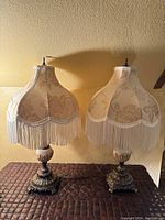 Two cream fabric lamps with floral shaded, each with long cream-colored fringe and ornate metal and ceramic bases