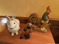 Photo showing a life size white rabbit figurine, a colorful metal rooster napkin holder, and a wooden cat figurine in a fishing pose on a wooden surface.