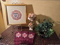 Photo showing all bathroom decor items together on brown textured surface: framed picture on gold stand, floral arrangement in vase, faux plant, and three embroidered washcloths.