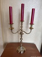 Full view of brass 3-tier candelabra with three red pillar candles on wooden surface against wallpapered wall.