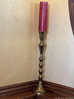 Full view of tall brass candle holder with red pillar candle set against beige wall and wood trim.