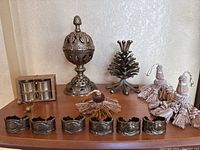 Photo of bronze hollow figurine, bronze pinecone figurine, boxed bronze napkin holders, bronze napkin rings, and tassels on table