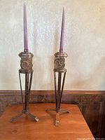 Two tall metal candle holders painted antique bronze, each with a purple taper candle inserted.