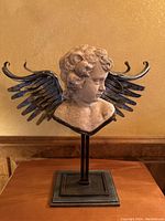 Front view of the plaster cherub bust with silver metal wings on a metal stand, showing detailed curly hair and the symmetrical wings.