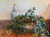 Overall view showing the tall white and gold decorative vase with lid alongside the planter with artificial ivy plant with trailing vines.