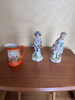 Photo showing two porcelain figurines, one of a girl with a goose and one of a boy, and a cream-colored ceramic pourable jar with orange handle and painted horse and cart scene.