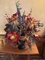 Front view of artificial flower arrangement with colorful flowers and pinecones in black metal vase placed on wooden surface.