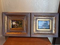 Two framed landscape paintings side by side on a flat surface, showing ornate deep gold and silver patinated wooden frames.