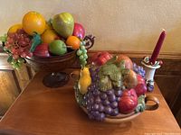 Overall view showing all three items: the plastic fruit bowl with metal base and artificial fruit, ceramic fruit bowl with fruit designs, and red candle on decorative holder.
