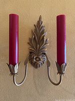 Gold painted metal wall sconce with two red candles mounted on wall, showing detailed leaf and flower design base.