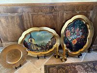 Lot showing two large ornate gold-painted plastic platters with floral and town paintings, a gold-painted terracotta charger plate, and two brass candle holders with yellow candles on tile floor against a wooden backdrop.
