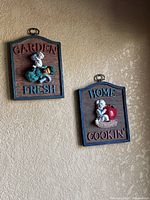 Two kitchen wall plaques mounted on a beige wall. One reads 'GARDEN FRESH' with a 3D boy figure holding vegetables. The other reads 'HOME COOKIN' with a boy figure holding an apple. Both plaques have a hanging loop at top.