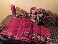 Whole set of maroon and gold hand towels and washcloths on a brown patterned surface with decorative boxes and artificial fruit centerpiece in background
