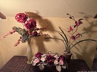 Three red floral arrangements displayed on a table against a wall: one in frosted glass vase, one in basket base, one red orchid in gold vase and stand.