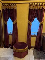 View of two red velvet curtains with gold tassel trim and a red velvet round vanity bench with gold tassel trim in between.
