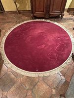 Photo showing a red/maroon circular rug with a cream fringe around the edge, approximately 92 inches diameter, placed on a tiled floor with some signs of use.