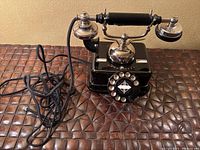 Front view of the black antique style rotary phone showing the rotary dial with letters and numbers, handset resting on cradle with silver metallic details, and black coiled cord.