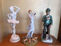 Front view of three figurine statues: white ballerina on the left, woman with puppies in center, old man with cane and dog on the right.