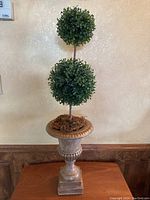 Full view of the artificial topiary tree with two green foliage spheres and classical urn planter on a wooden surface against textured wall.