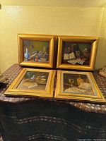 Four gold painted frames displayed together showing still life images with various objects such as musical instruments, books, and glassware.