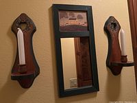 Two wooden sconces with white taper candles flanking a hunter green framed mirror mounted on a wall.