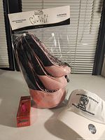 Set of three pink and black lace Glove It golf club headcovers, boxed Srixon pink Lady golf balls, and white embroidered hat arranged on a table.