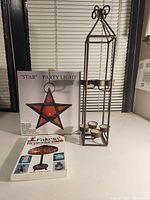 Photo of clear glass star hanging candle light with chain, metal candle holder stand, and book 'Fakes & Reproductions' on table.