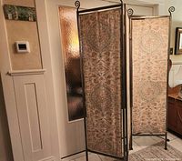 Three-panel metal frame room divider shown partially open, fabric pattern on front side includes detailed damask design in earth tone colors.