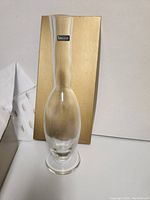 Tall clear glass vase or decanter on a white surface with a gold-colored box in the background. Shows the full shape and size of the item.