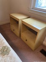 Two light wood finish nightstands placed side by side on a carpeted floor near a window, showing front and top surfaces.
